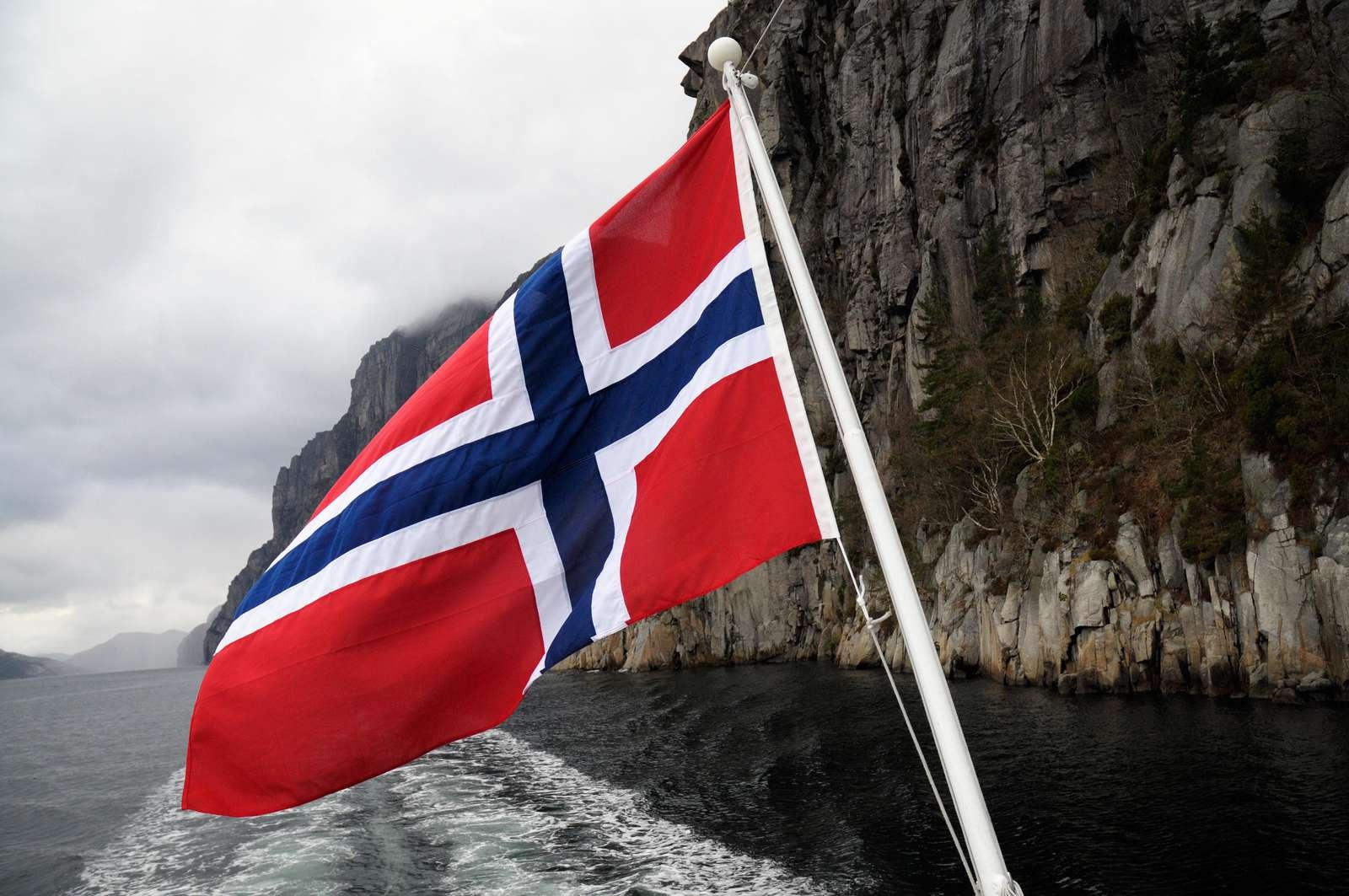 A boat is sailing with a Norwegian flag at the end of the boat. The rocks of Lysefjorden and a cloudy sky are in the background.